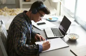 Jovem estudante concentrado estudando em casa com fones de ouvido, anotando em um caderno ao lado de um laptop