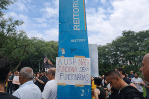 Protesto em frente à Reitoria da USP durante greve universitária, com manifestantes e cartaz sobre a paralisação dos funcionários.