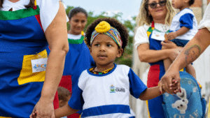 Criança com uniforme escolar da Prefeitura do Crato segura as mãos de adultos durante atividade de educação infantil.