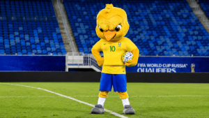 Mascote amarelo da Seleção Brasileira segura uma bola de futebol em campo, com arquibancadas azuis ao fundo.